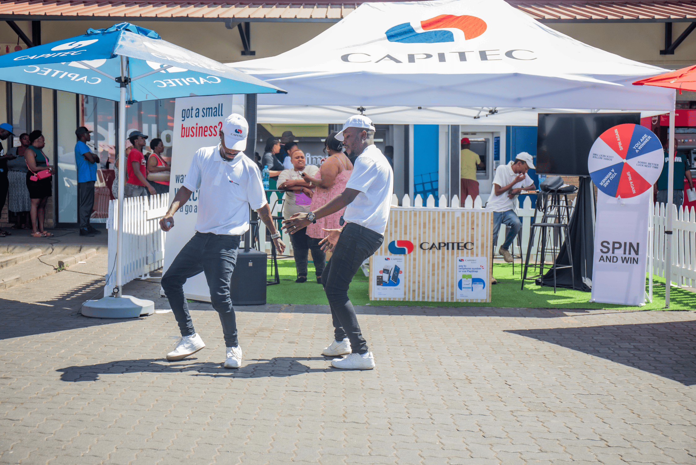 Two men in white shirts and hats walking past a Capitec bank tent and promotional stand.